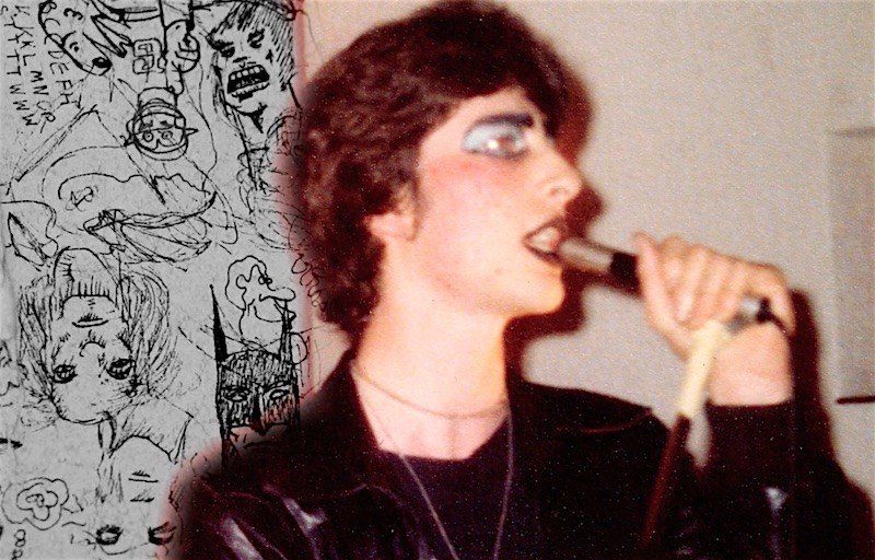 Photo of Neil Gaiman, holding a microphone. He is dressed all in black and wearing blue eyeshadow and black lipstick.